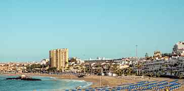 Sandstrand med mange blå parasoller, havet og bygninger i bakgrunnen. (AI-generert tekst)