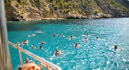 Svømmende mennesker i klart blått vann foran en klipperik kystlinje. (AI-generert tekst)