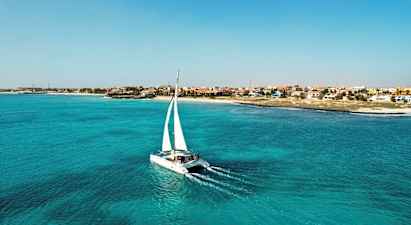 En katamaran med hvite seil seiler over turkis vann nær en kyst med hus i ulike farger. (AI-generert tekst)