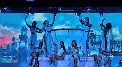 Dansere poserer på en opplyst scene med dekor av palasslignende bygninger mot en skyet himmel. (AI-generert tekst)