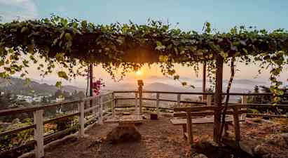 En utsiktspunkt med en trepergola dekket av blader samt benker og rekkverk rundt. Solnedgang over et fjellandskap i bakgrunnen. (AI-generert tekst)