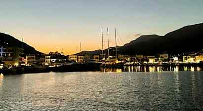 En havn i skumringen med båter fortøyd ved en kai og opplyste bygninger i bakgrunnen. Fjell kan sees i bakgrunnen. (AI-generert tekst)