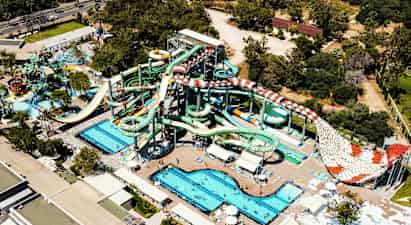 Et vannland med flere store vannsklier, bassenger og solsenger, omgitt av grønne trær og bygninger. (AI-generert tekst)