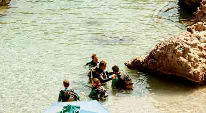 Gruppe personer i våtdrakter og dykkerutstyr står i klart vann ved steiner. (AI-generert tekst)