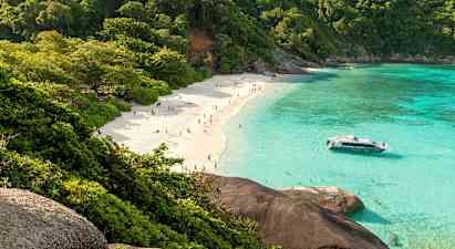 En sandstrand med mennesker, omgitt av frodige trær og klart turkis vann. En båt ligger nær stranden. (AI-generert tekst)