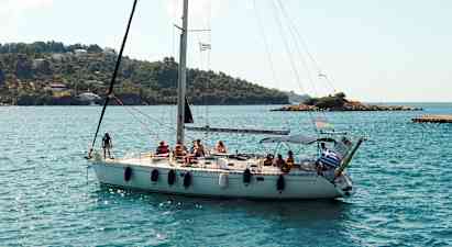 En seilbåt med flere personer om bord seiler nær en kystlinje med grønne åser og noen hus. (AI-generert tekst)