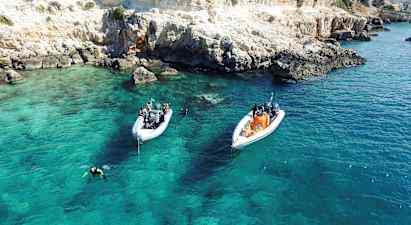 To båter ligger for anker i klart turkist vann nær en klippefull kyst. Dykkere er i vannet. (AI-generert tekst)