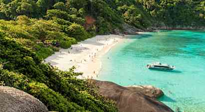 En sandstrand med mennesker, omgitt av frodige trær og klart turkis vann. En båt ligger nær stranden. (AI-generert tekst)