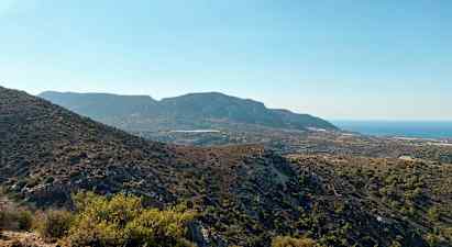 En utsikt over et fjelllandskap med variert vegetasjon og havet i bakgrunnen under en klar blå himmel. (AI-generert tekst)
