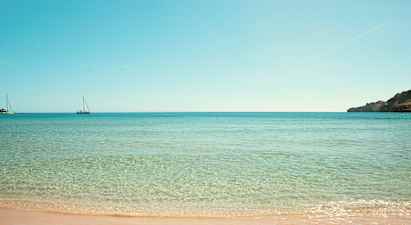 Strand med klart vann og to seilbåter i havet. (AI-generert tekst)