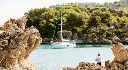 En seilbåt ligger fortøyd ved en fredelig vik omgitt av klipper og skog, med en person som fisker fra stranden. (AI-generert tekst)