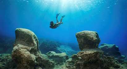 En snorkler svømmer under vann mellom store steiner med sollys som filtrerer ned fra overflaten. (AI-generert tekst)