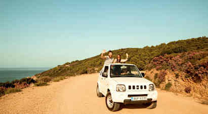 En hvit bil parkert på en grusvei ved siden av en kystlinje med skogkledde åser. (AI-generert tekst)