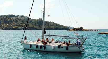 En seilbåt med flere personer om bord seiler nær en kystlinje med grønne åser og noen hus. (AI-generert tekst)