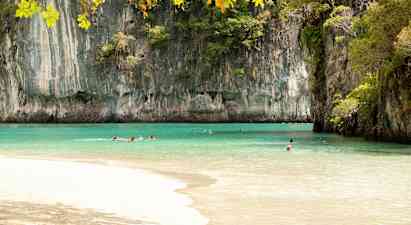 Bildet viser en strand med klart tyrkis vann omgitt av høye klippeformasjoner dekket av vegetasjon. Flere personer svømmer i vannet under en klar himmel. (AI-generert tekst)
