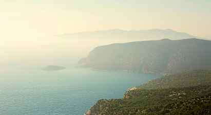 En kystlinje med bratte klipper og grønne skoger som skråner ned mot havet. Et dis dekker landskapet. (AI-generert tekst)