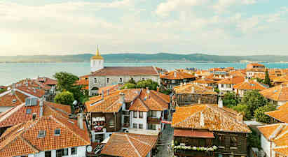 Gamlebyens tak med hus med røde tegltak og en kirke i bakgrunnen nær havet ved kysten. (AI-generert tekst)