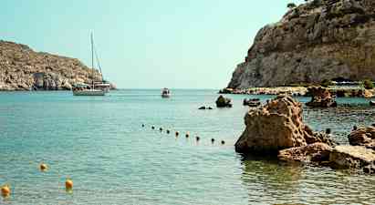 En bukt med klart vann og steinete kyst, hvor en båt og en mindre flytende fartøy er synlige i bakgrunnen. (AI-generert tekst)