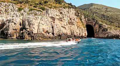 En motorbåt med folk kjører på blått vann nær en klippe med en stor hulåpning. (AI-generert tekst)
