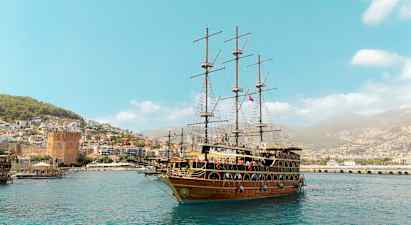 Et stort seilskip ligger ved havnen, med en by og grønne åser i bakgrunnen. (AI-generert tekst)