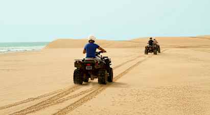 To personer kjører firehjulinger over en sandstrand med havet i bakgrunnen. (AI-generert tekst)