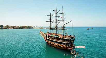 Et stort treskip med seil som ligger i rolig vann, omgitt av svømmere, og i bakgrunnen sees kystlinjen. (AI-generert tekst)