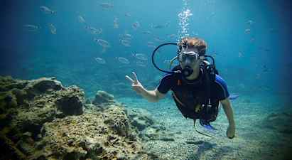 En person i dykedrakt under vann omgitt av små fisker og med en steinet havbunn i bakgrunnen. (AI-generert tekst)