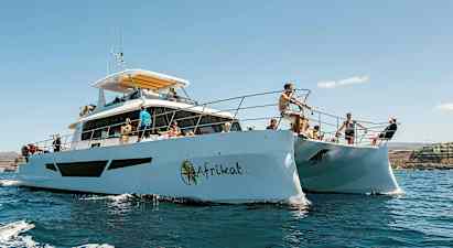 Personer på en katamaran i solfylt vær, omgitt av vann og en kystlinje i bakgrunnen. (AI-generert tekst)