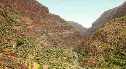 En smal vei slynger seg gjennom en dal omgitt av bratte fjell med grønn vegetasjon. (AI-generert tekst)