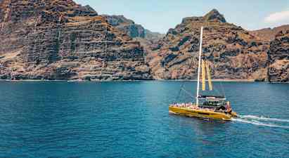 En katamaran seiler nær høye klipper, med klart blått vann i forgrunnen. (AI-generert tekst)