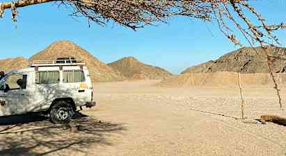 En hvit bil står parkert under et tre i en ørkenomgivelse med fjell i bakgrunnen. (AI-generert tekst)