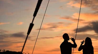 Silhuetter av to personer som holder glass ved solnedgang med havet og en liten øy i bakgrunnen. (AI-generert tekst)