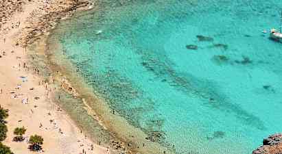 Strand med gyllen sand og klart turkis vann. Mange mennesker nyter solen og svømmer. En seilbåt ligger for anker lenger ute. (AI-generert tekst)