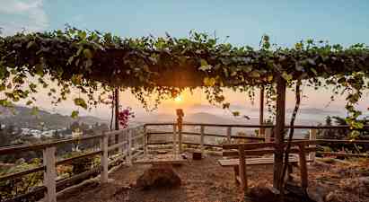 En utsiktspunkt med en trepergola dekket av blader samt benker og rekkverk rundt. Solnedgang over et fjellandskap i bakgrunnen. (AI-generert tekst)