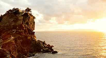 En klippe ved kysten med en bygning og trær på toppen. Havet sees under en solnedgang. (AI-generert tekst)