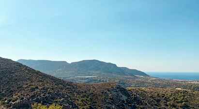 En utsikt over et fjelllandskap med variert vegetasjon og havet i bakgrunnen under en klar blå himmel. (AI-generert tekst)