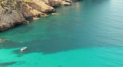 En båt ligger ankrert i klart turkis vann nær en steinete kystlinje med klipper. (AI-generert tekst)