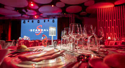 En borddekking med vinglass i et rom opplyst med røde lys. Bakgrunn med en stor skjerm med teksten 'Scandal' og en scene. (AI-generert tekst)