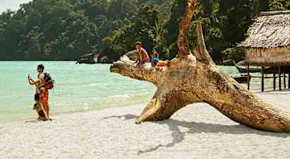 To barn sitter på en stor trestokk på en sandstrand ved havet, omgitt av frodig skog og trebygninger. (AI-generert tekst)