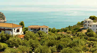 Et landskap med hvite hus og røde tak omgitt av grønn vegetasjon og utsikt over et rolig blått hav. (AI-generert tekst)