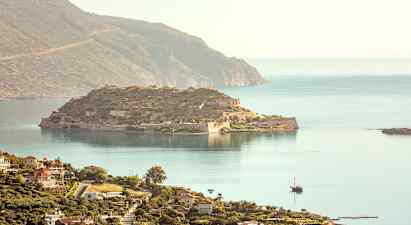 En utsikt over en historisk øy med ruiner og omkringliggende hav, og i forgrunnen sees en kystby med bygninger. (AI-generert tekst)