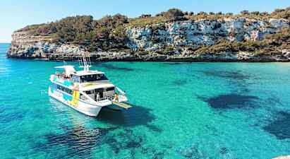 En hvit båt med passasjerer i en turkisblå vik, omgitt av klipper og grøntområder. (AI-generert tekst)