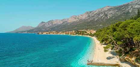 Utsikt over en rullesteinstrand med turkist vann og frodige trær. Fjell og en landsby vises i bakgrunnen. (AI-generert tekst)
