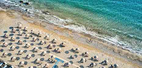 Luftfoto av en sandstrand med solsenger og parasoller, med kystlinjen og blått hav i bakgrunnen. (AI-generert tekst)