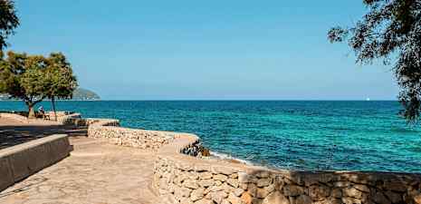En steinmur ved havet med blå himmel og synlige trær i forgrunnen. (AI-generert tekst)