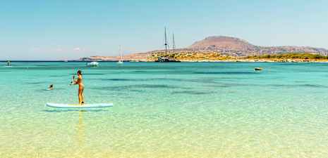 Person som står på et paddleboard i klart blått vann med en fjellrik kystlinje i bakgrunnen. (AI-generert tekst)