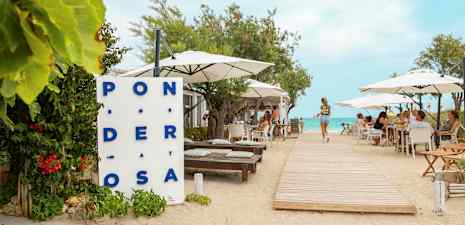 En restaurant ved stranden med bord og parasoller, en tresti fører mot vannet. (AI-generert tekst)