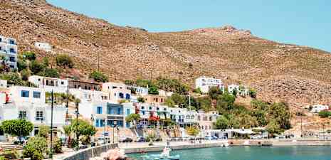 En kystby med hvite og fargerike bygninger, små båter ved en steinete kystlinje og tørre fjell i bakgrunnen. (AI-generert tekst)