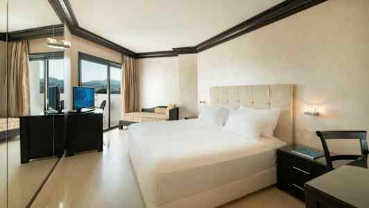 Hotellrom med dobbeltseng, skrivebord med stol og TV samt en stor vindusdør. (AI-generert tekst)