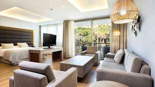 Et hotellrom, dekorativt møblert med sofa, lenestoler, seng og TV, samt utsikt mot en frodig have. (AI-generert tekst)